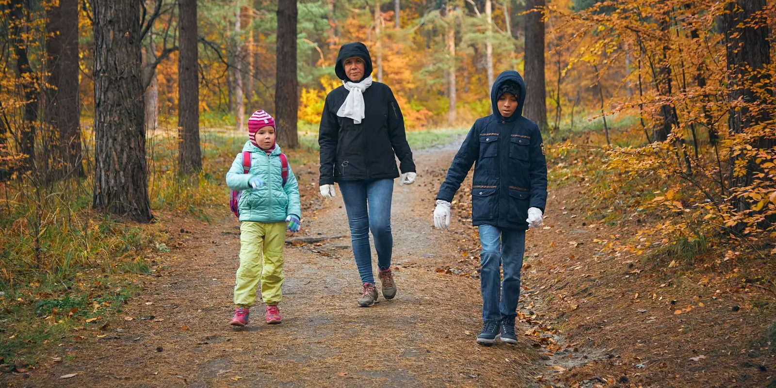 A woman and two children in warm coats walk along a woodland path in autumn