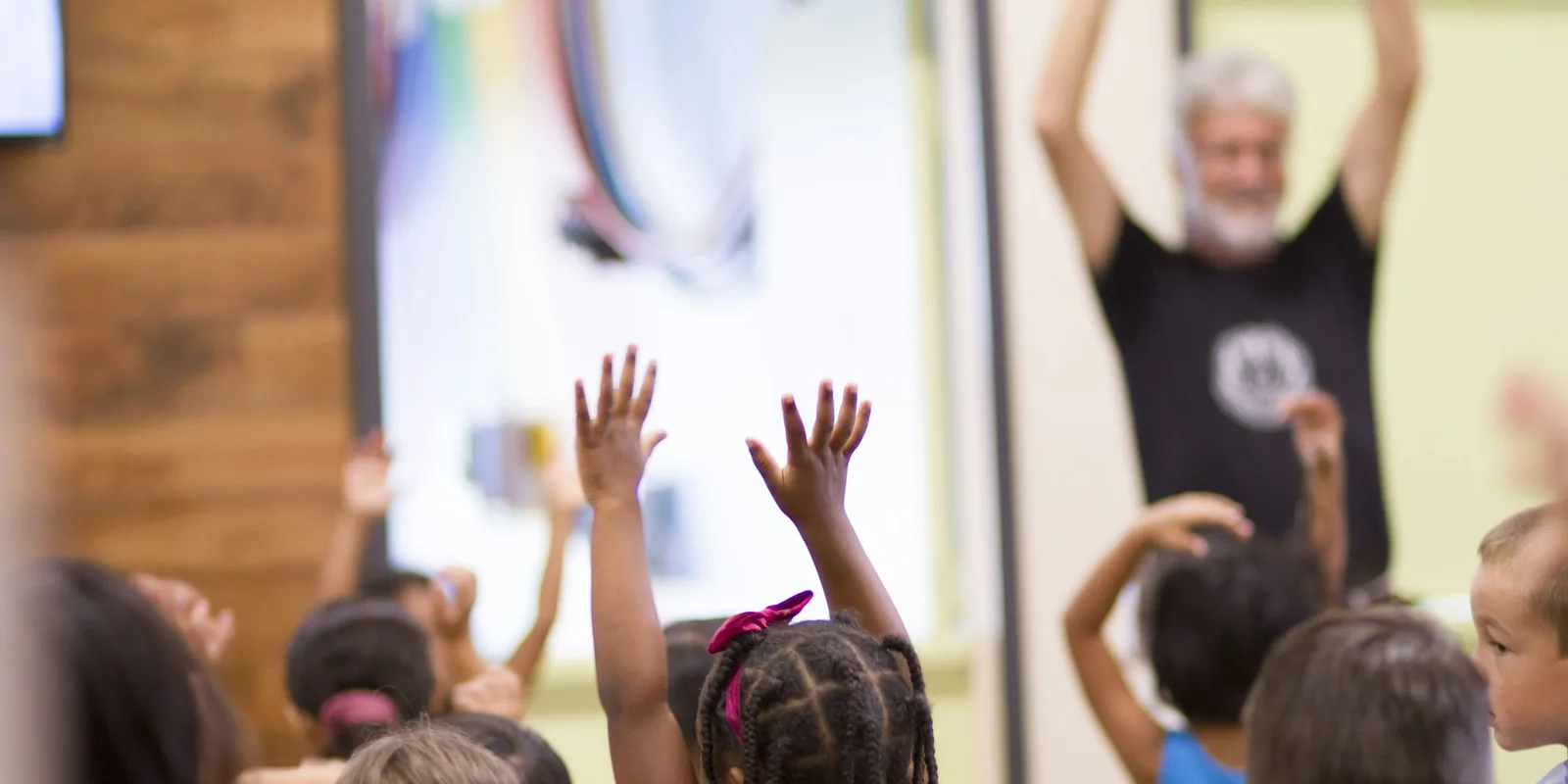 Children raise their arms in the air copying a man leading a song at the front of the room