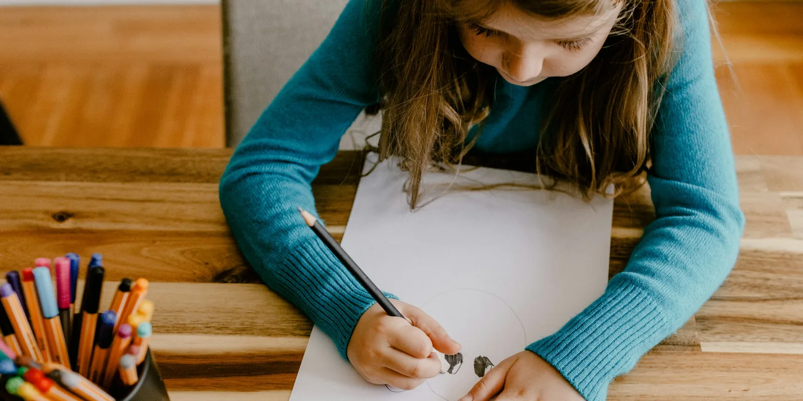 A girl in a blue jumper draws with a pencil at a wooden table, a pot of felt-tip pens beside her
