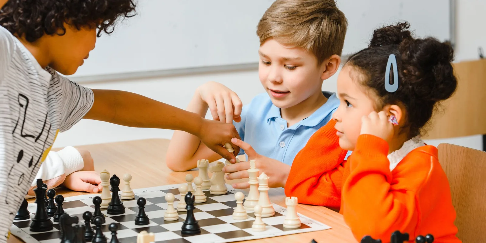 Three children play chess at a classroom table, one reaching across to move a piece