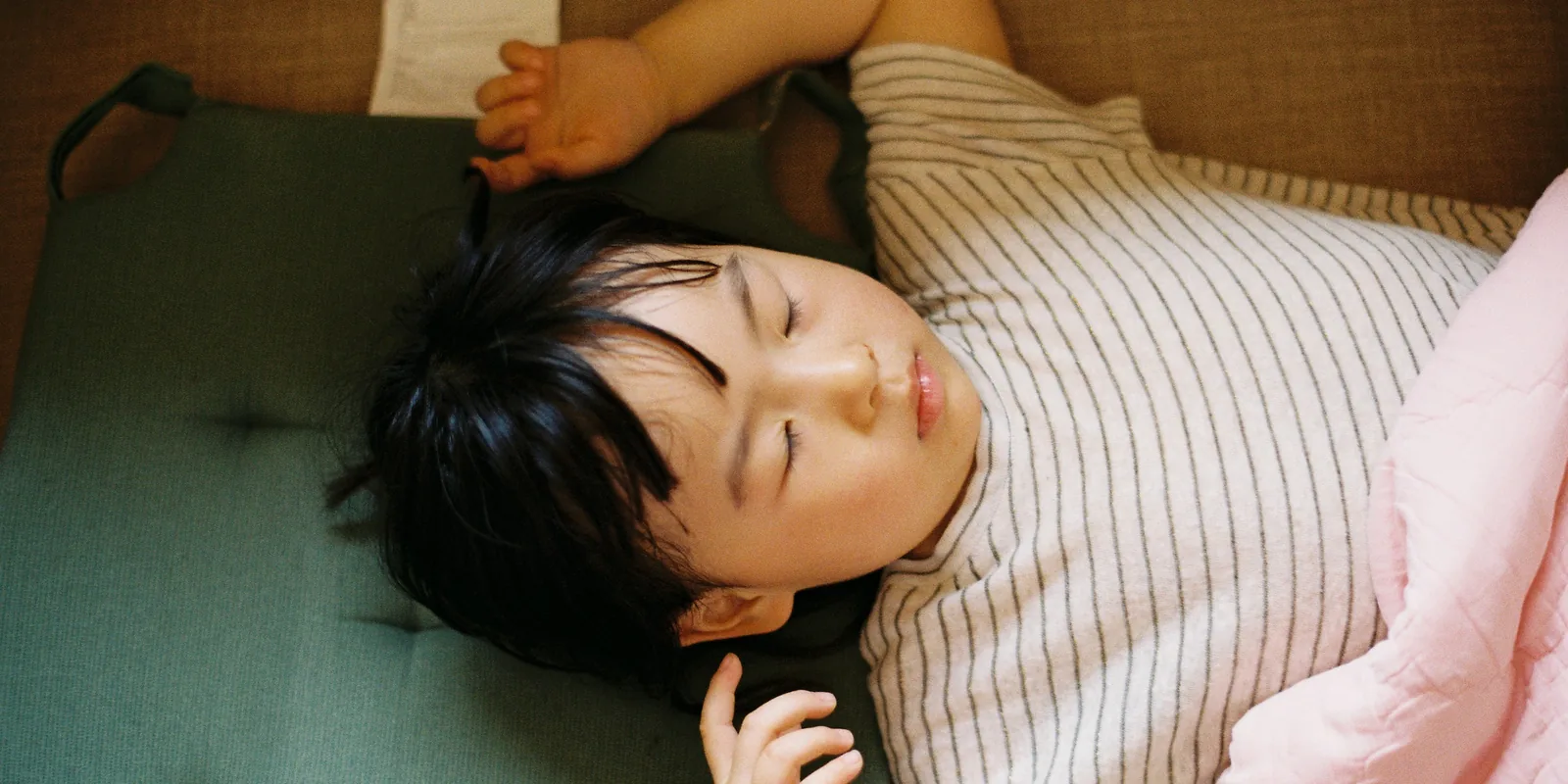 A young child in a striped top lies on a cushion with eyes closed, arms flung out, worn out