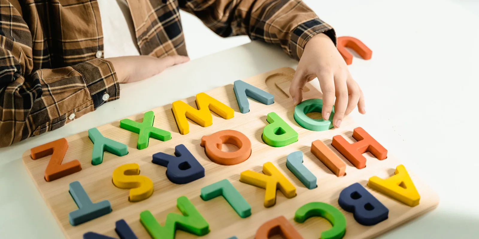 A child fits coloured wooden letters into an alphabet puzzle board on a white table