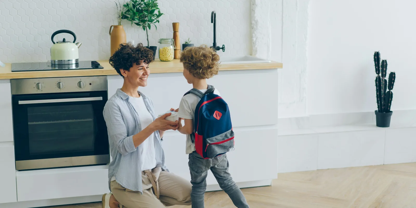 A smiling woman kneels in a kitchen to hand a lunchbox to a boy wearing his school rucksack