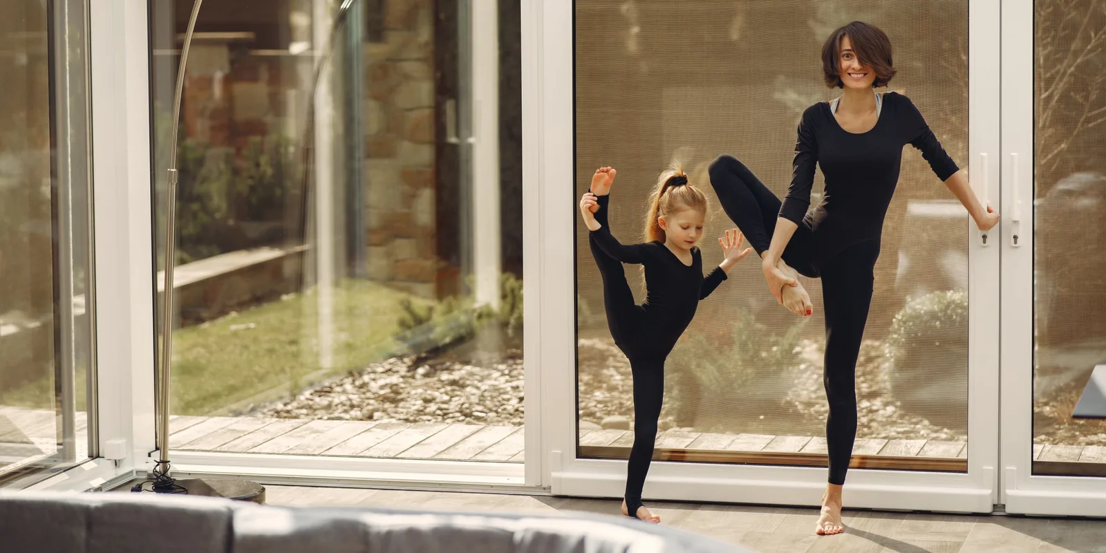 A girl and a woman each balance on one leg with the other foot held high, by a glass door at home