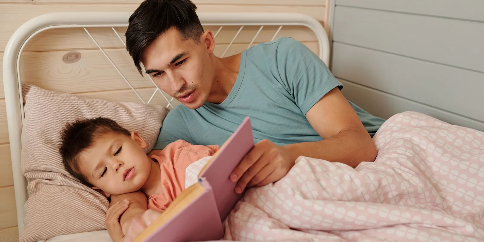 A man lies beside a small boy in bed, reading him a story from a pink book