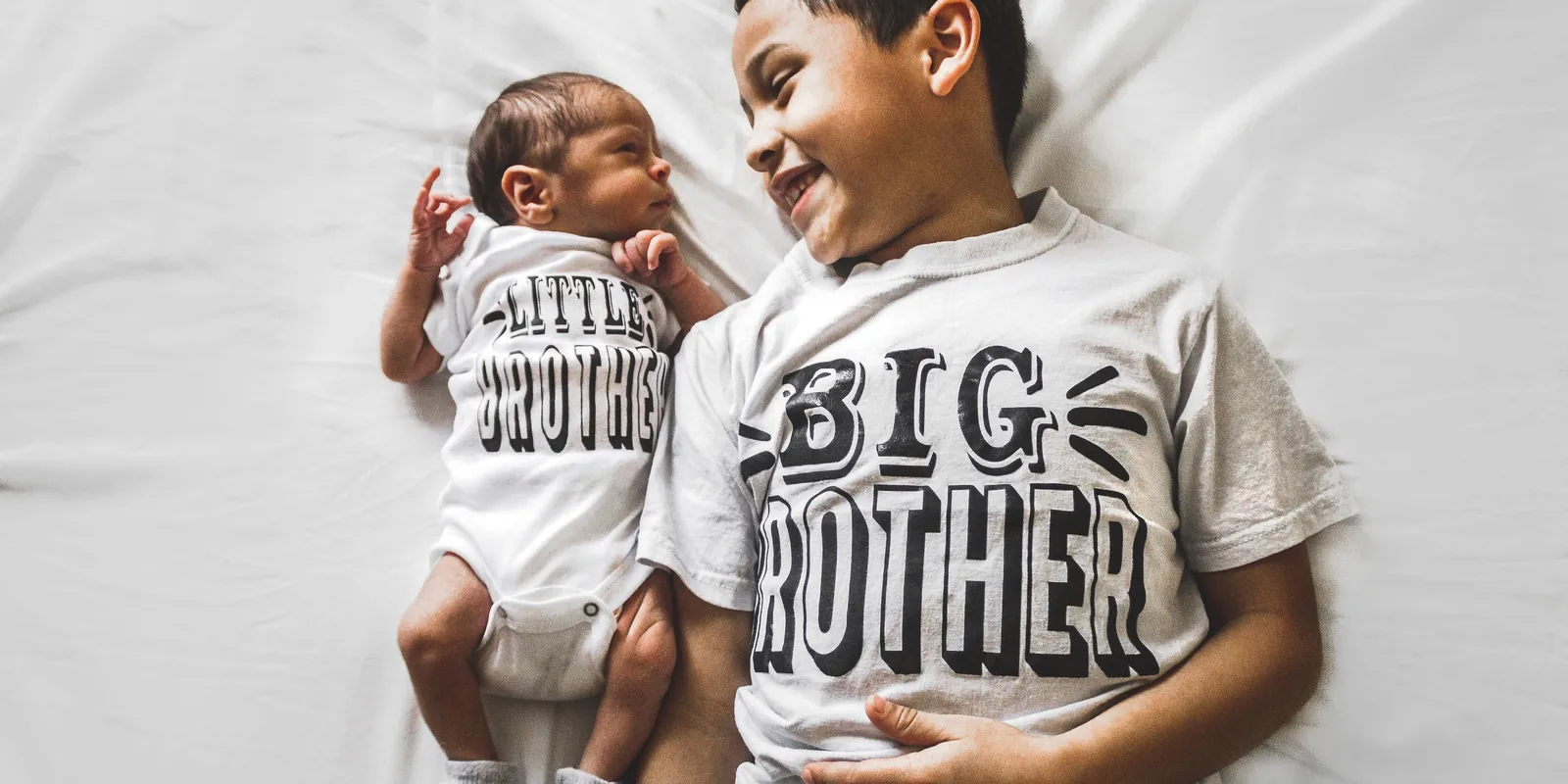 A smiling boy in a Big Brother T-shirt lies next to a newborn in a matching Little Brother vest