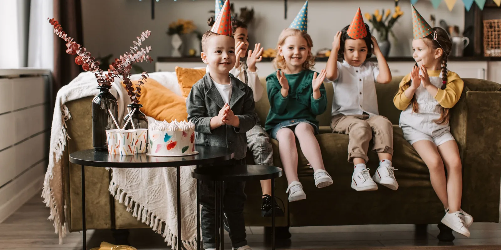 Five children in paper party hats sit on a sofa clapping, with a cake on the table beside them