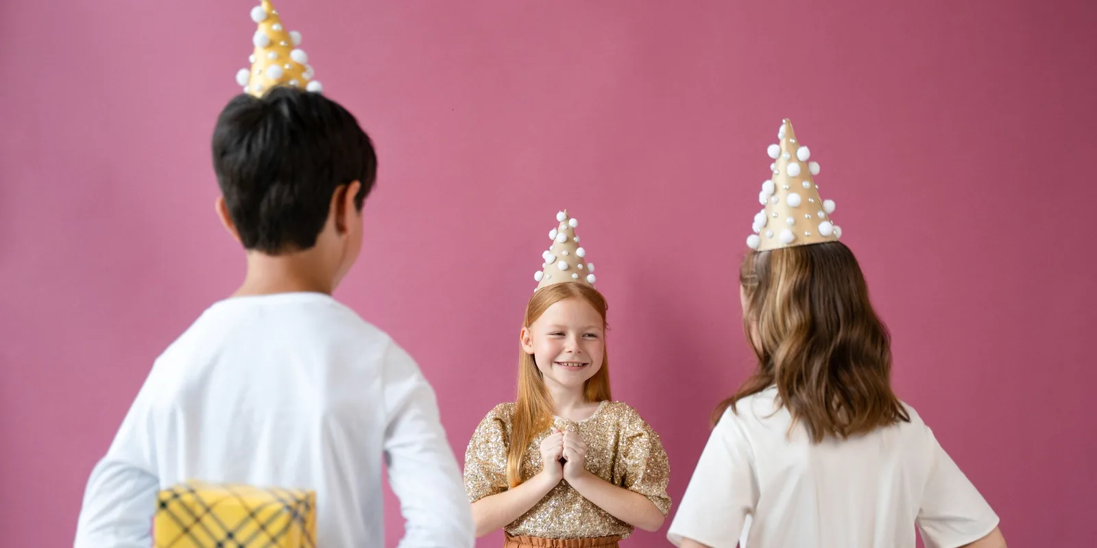 Two children in party hats hide wrapped presents behind their backs as a smiling girl in a gold top waits for them