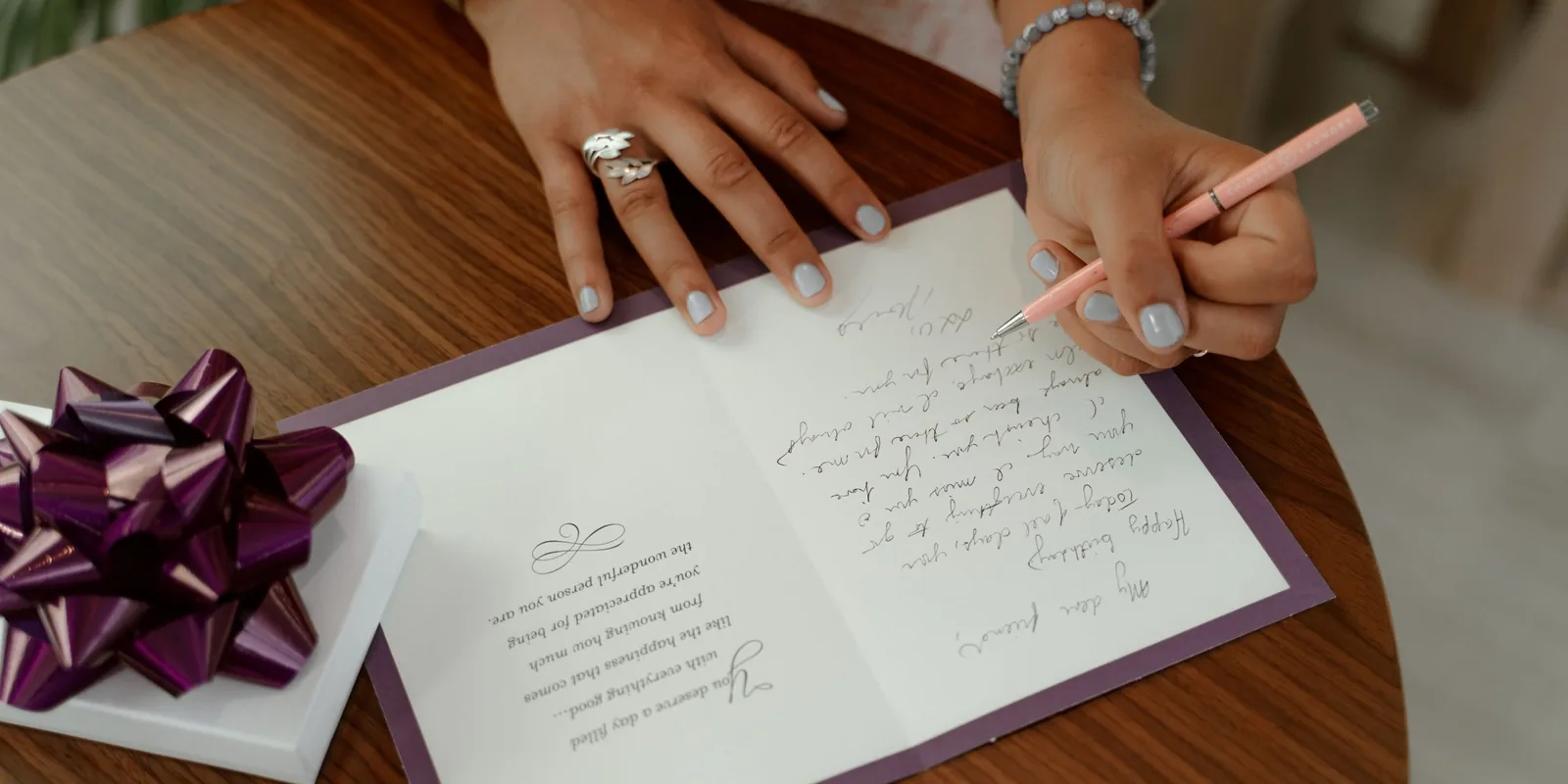A woman writes a handwritten birthday message inside a card, next to a gift topped with a purple bow