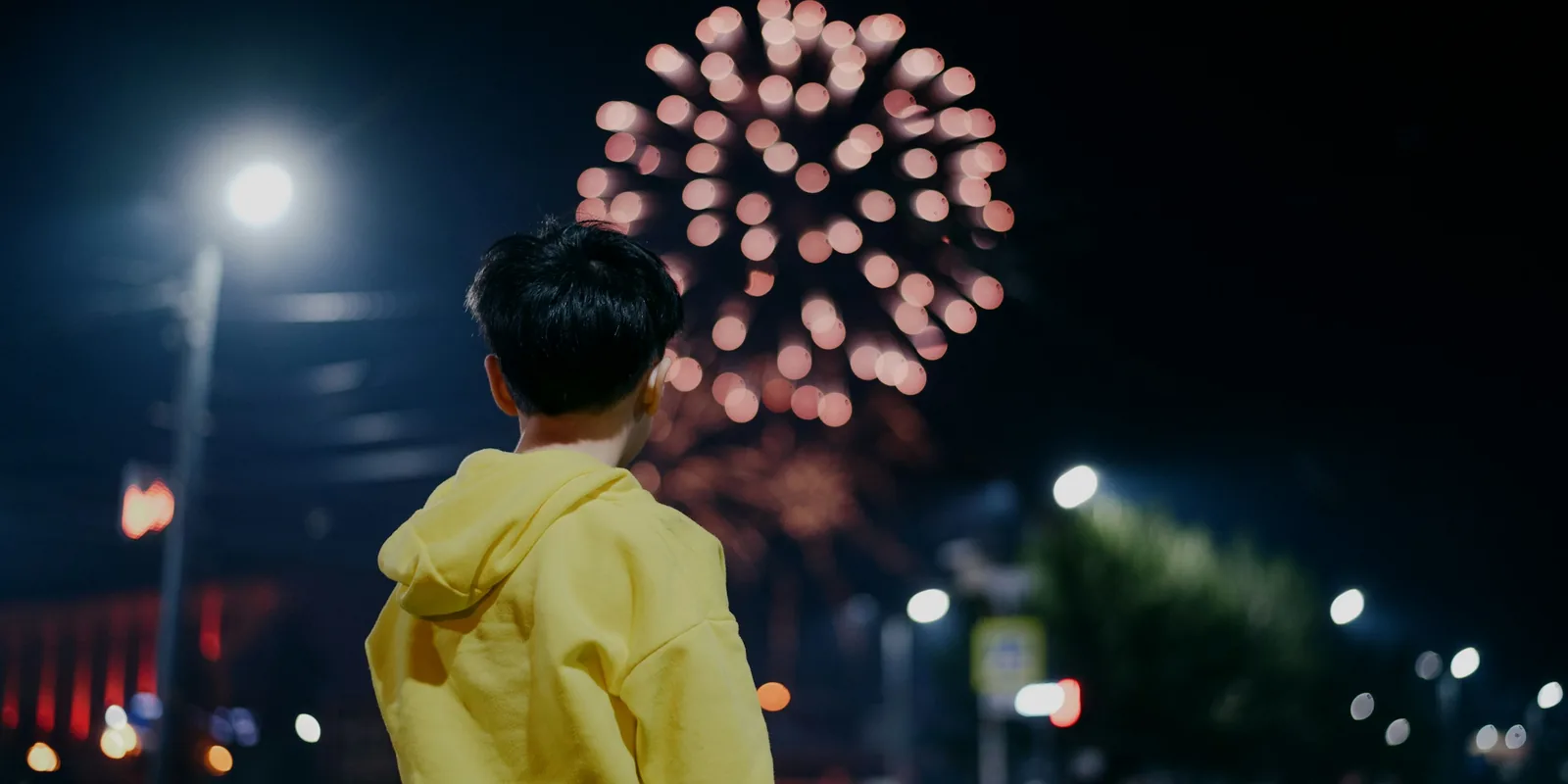 A boy in a yellow hoodie watches a firework burst over a lamp-lit street at night