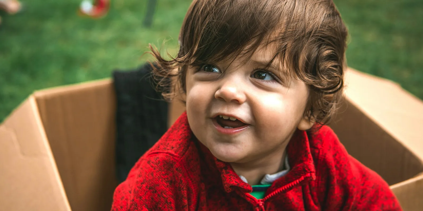 A smiling toddler in a red fleece sits inside a cardboard box on the grass