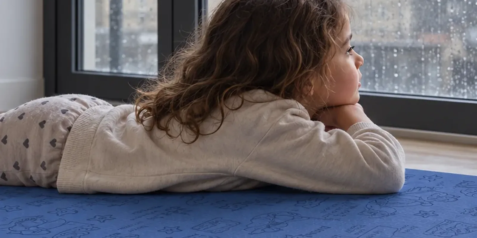 A child lies on her front on a blue Yogi-Me mat, chin resting on her hands, watching rain run down the window