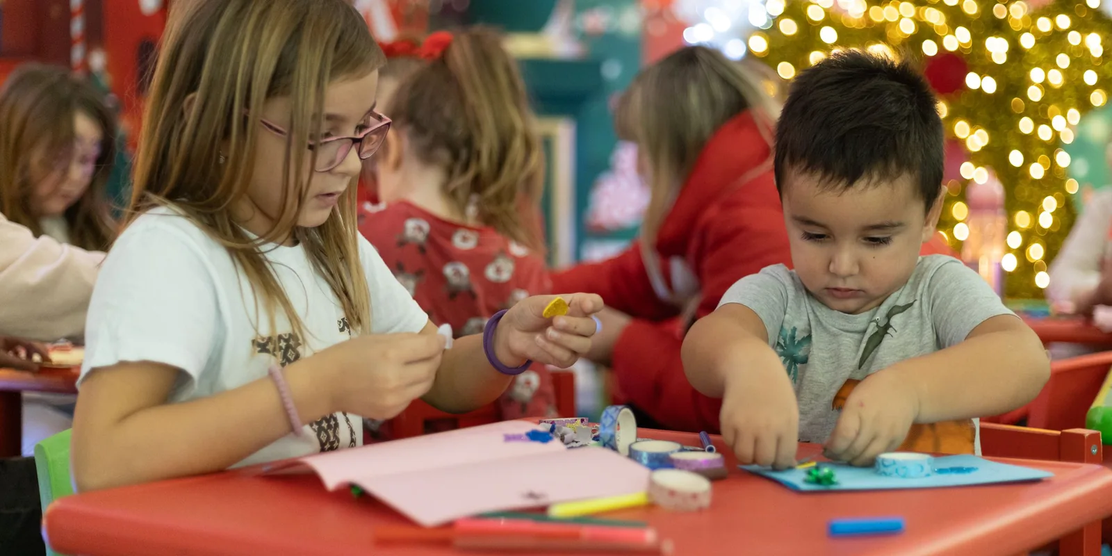 A girl and a younger boy make paper crafts at a red table in a classroom decorated for Christmas