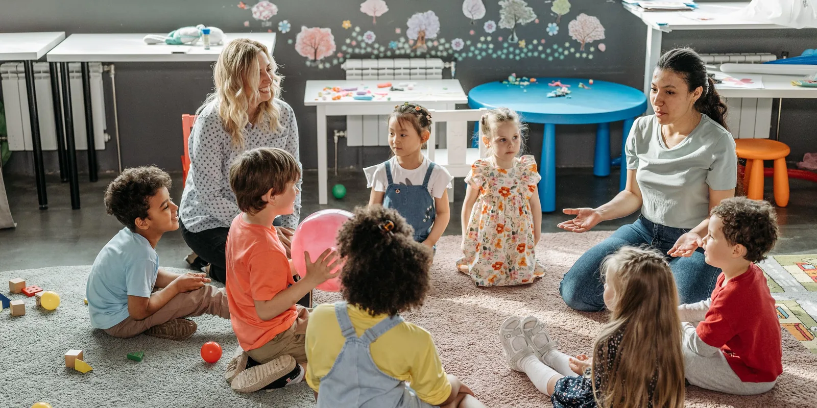 Nursery children sit in a circle on the carpet with two teachers, passing a pink ball