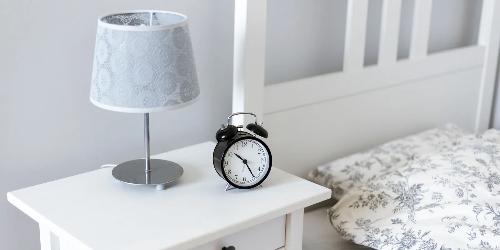 A black alarm clock and a small lamp sit on a white bedside table next to a bed with a floral pillowcase
