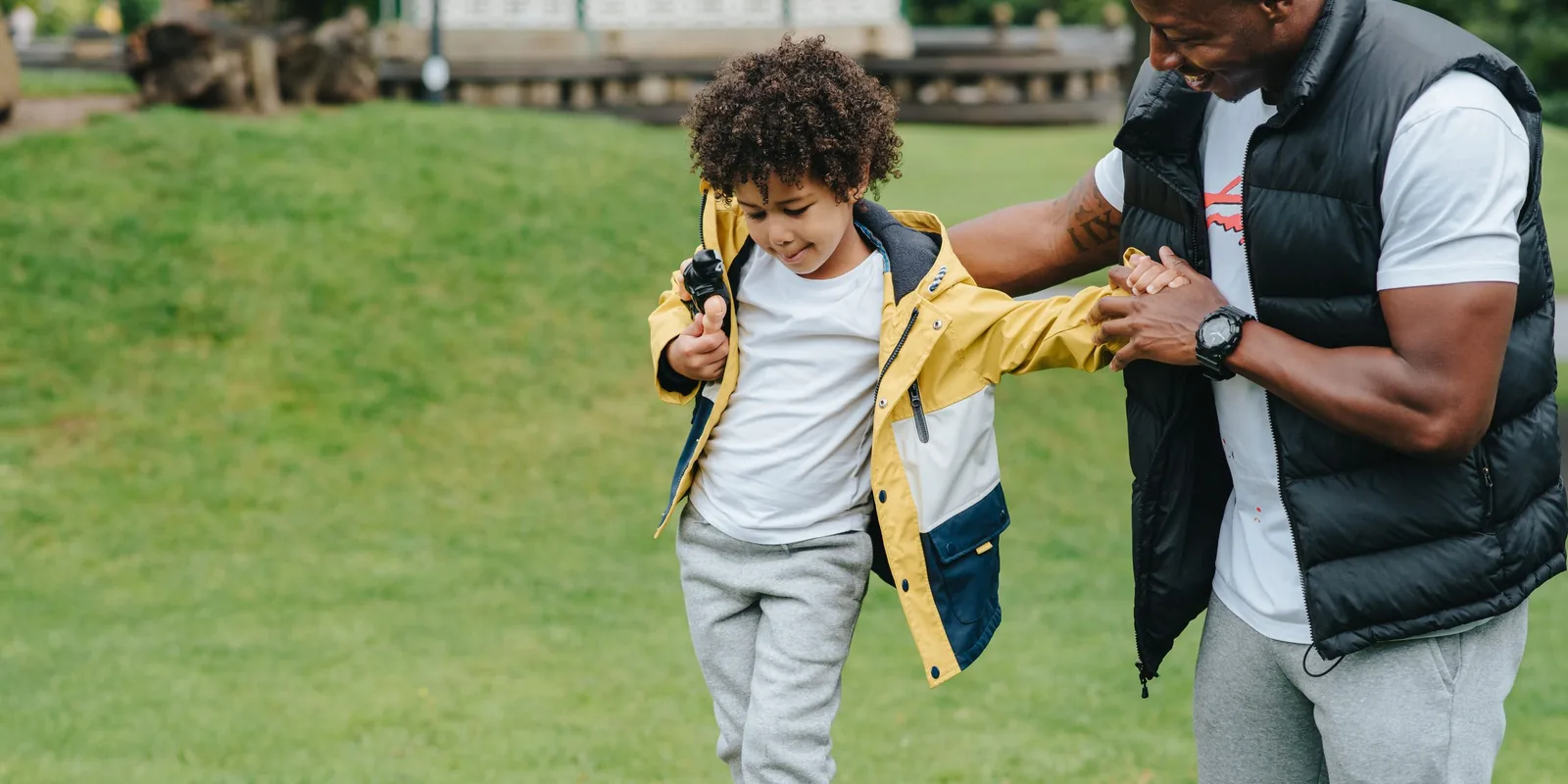 A boy in a yellow raincoat balances along a fallen log in a park while an adult steadies his arm