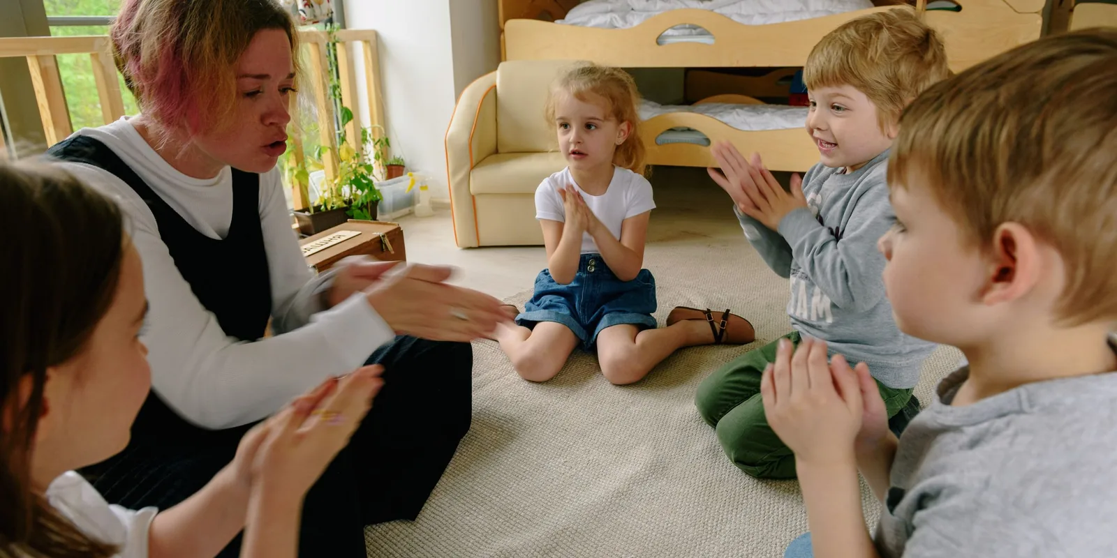 A nursery teacher leads a clapping game with young children sitting in a circle on the floor