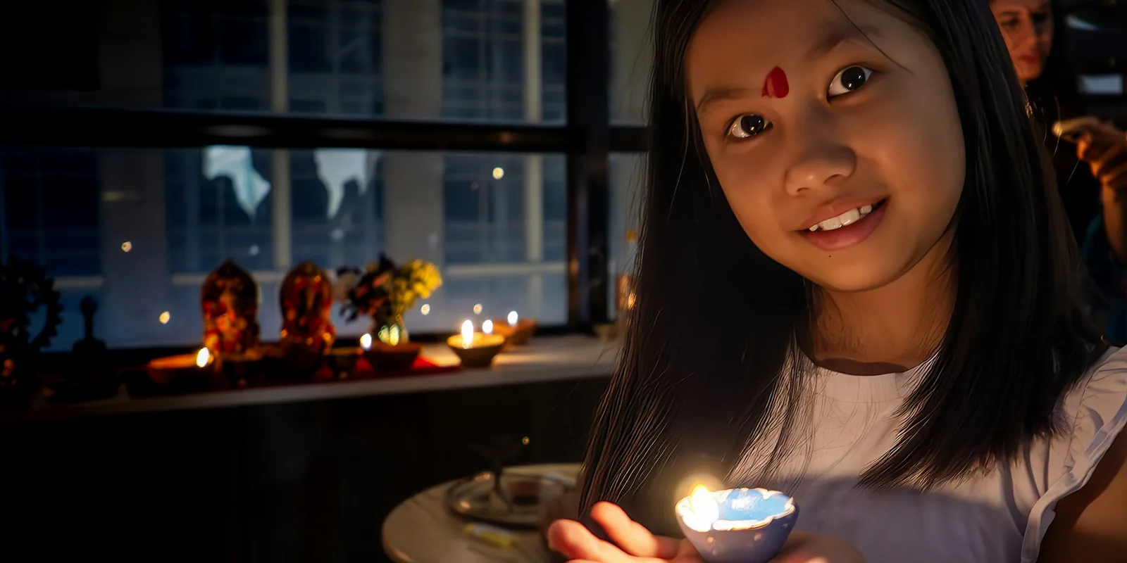 A smiling girl holds a lit clay diya in her palm, with more lamps glowing on the windowsill behind her