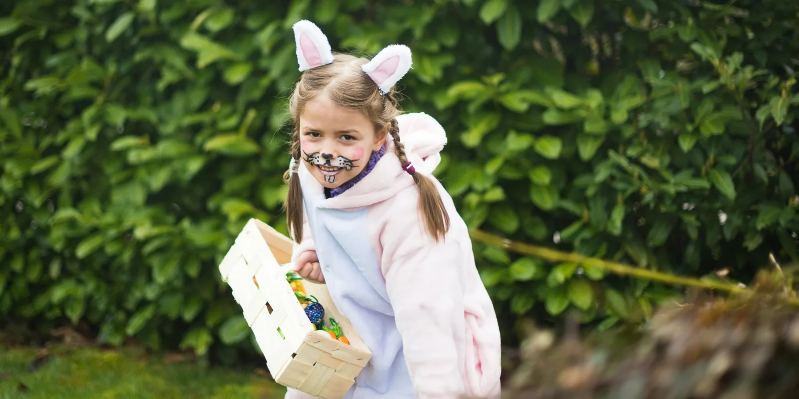 A girl with bunny ears and painted whiskers carries a small wooden basket of Easter eggs past a hedge