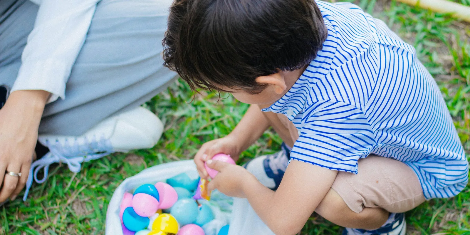 A boy crouches on the grass opening a pink plastic egg from a bag of eggs he has found