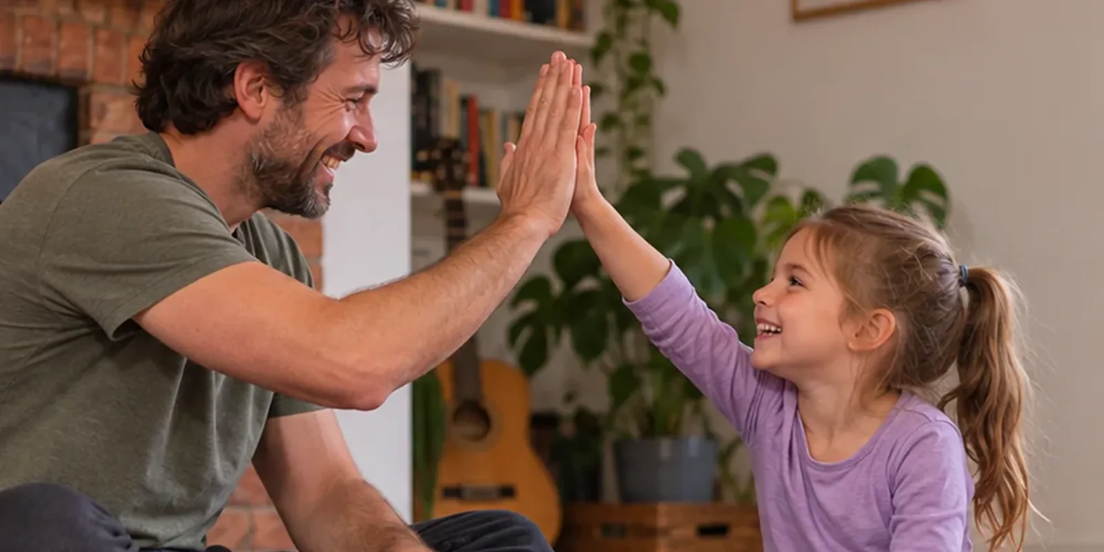 An adult and a young girl sit on the floor of a living room, laughing as they share a high five