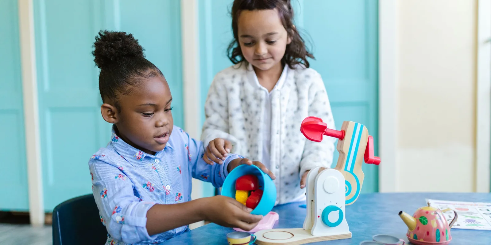 Two girls play with a wooden toy food mixer and a tin teapot at a classroom table