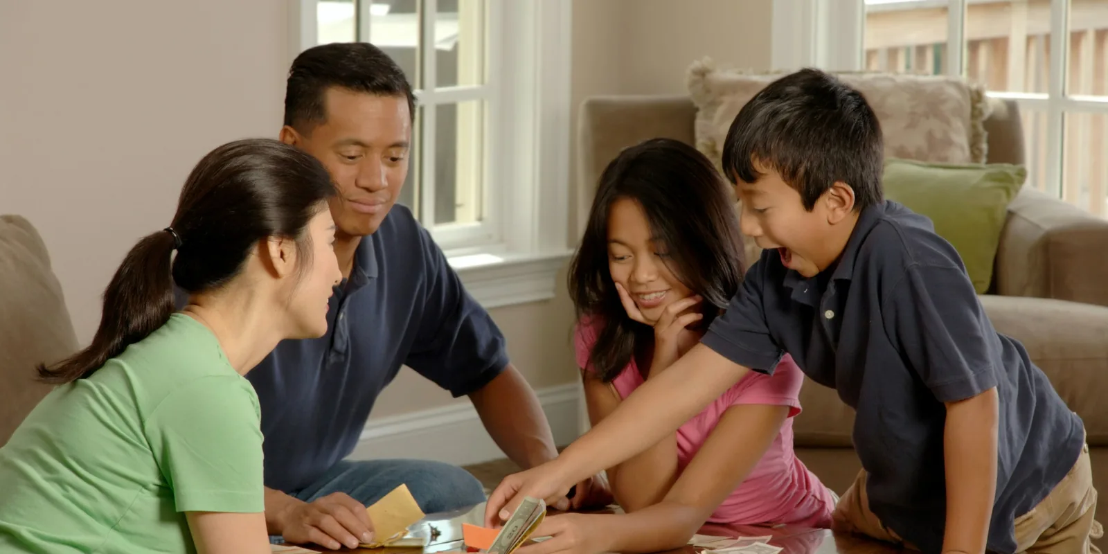 Two adults and two children gather round a coffee table mid-game, the boy reaching excitedly for the play money