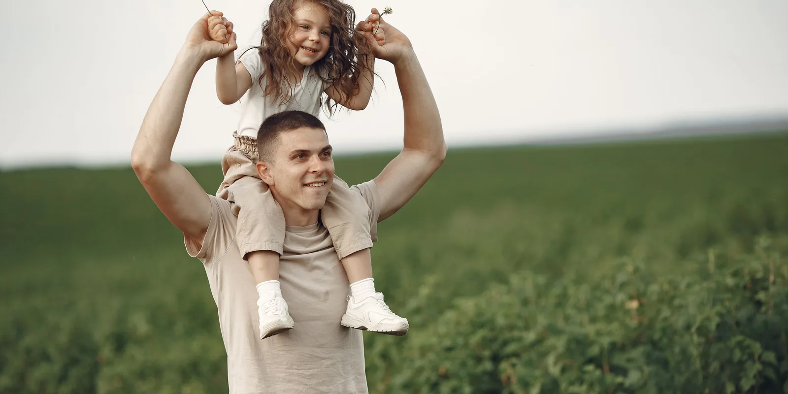 A smiling man carries a little girl on his shoulders across a green field, holding her hands up high