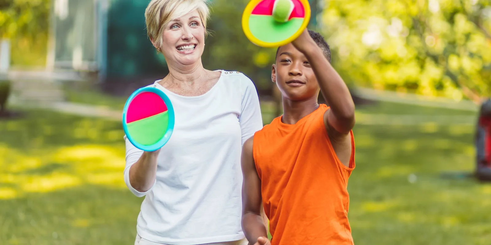 A boy and a woman play a catch game with bright velcro paddles on a lawn