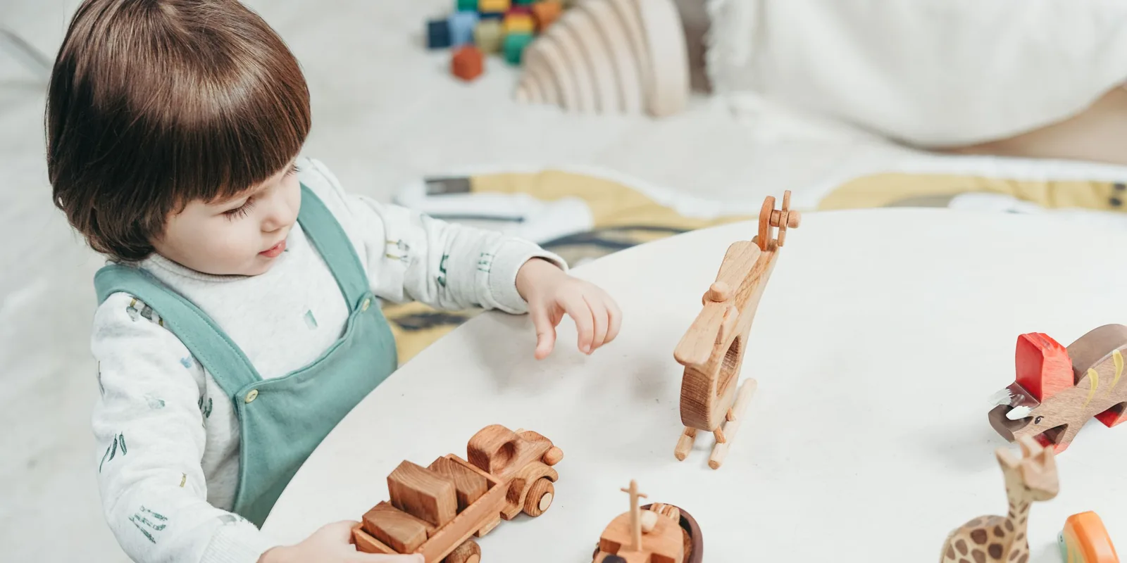 A toddler in green dungarees plays with wooden toy vehicles and animals on a white table