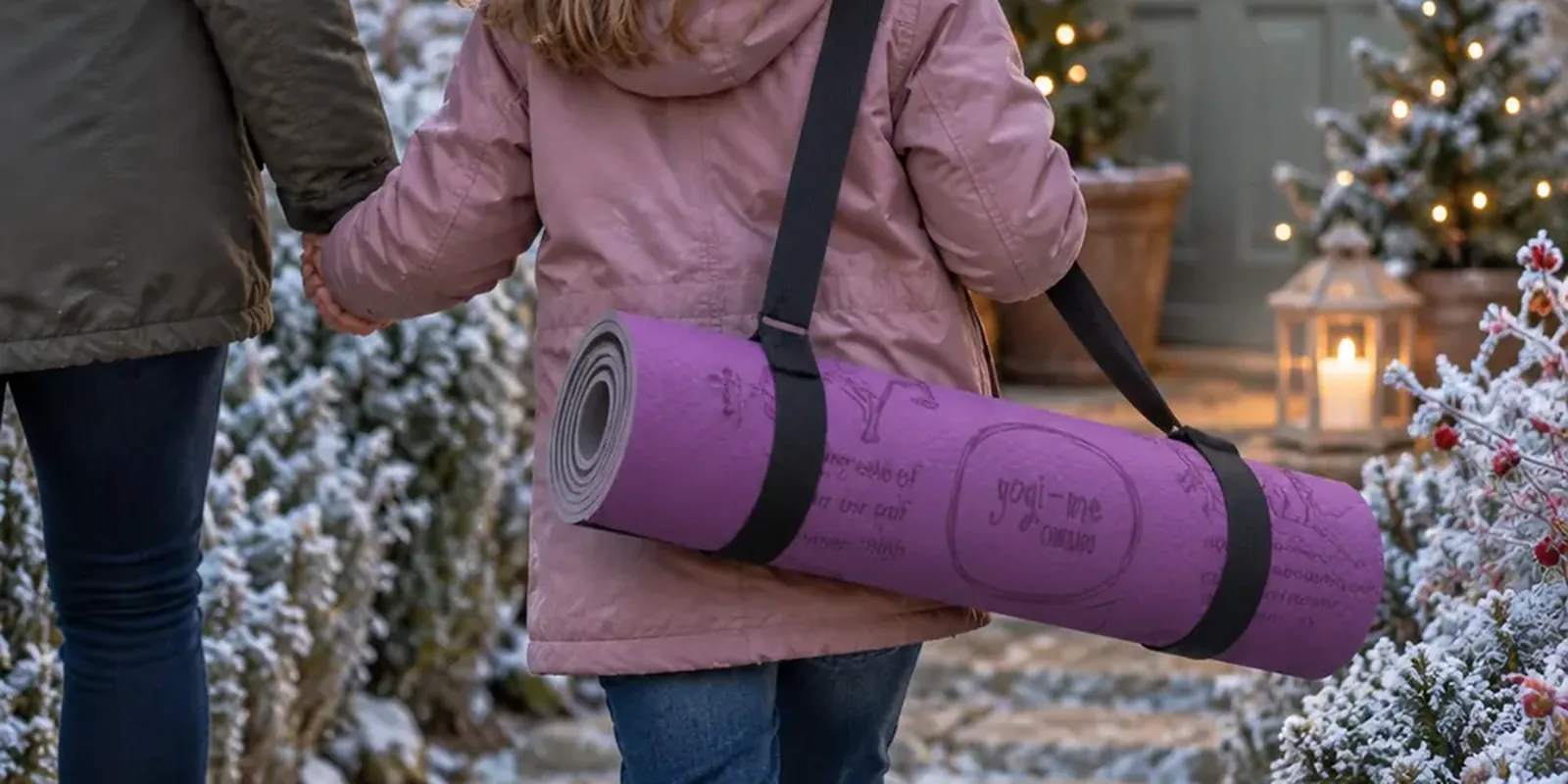 A child in a pink winter coat walks up a frosty path holding an adult's hand, a purple Yogi-Me mat on its carry strap across her back