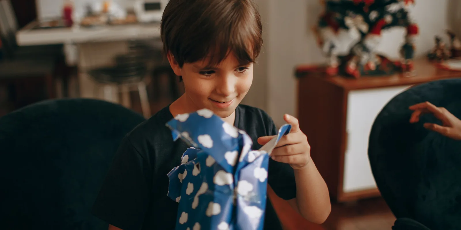 A boy smiles as he tears open a present wrapped in blue paper with white clouds