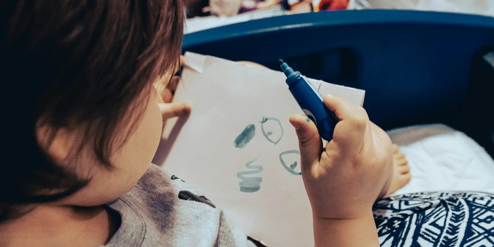 A young child holds up a sheet of paper and a blue felt-tip, drawing a face