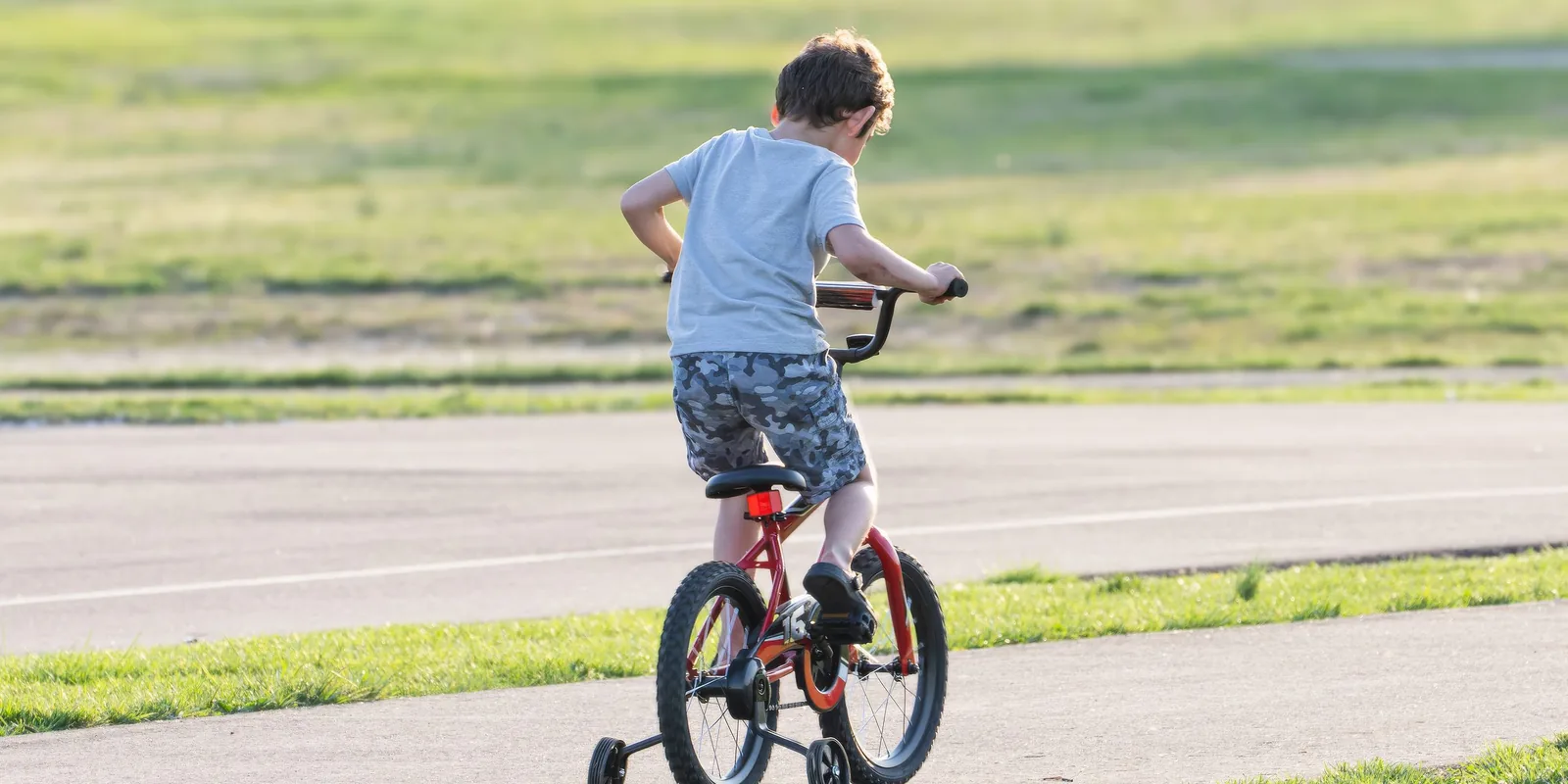 A boy pedals a small red bike with stabilisers along a path beside a grassy field