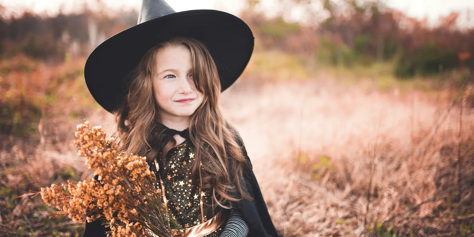 A smiling girl in a witch's hat and starry cape holds a bunch of dried flowers in an autumn field