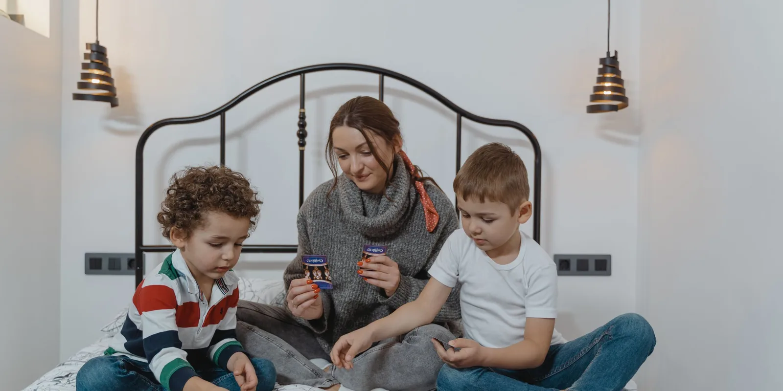 A mother and two boys sit cross-legged on a bed playing a card game