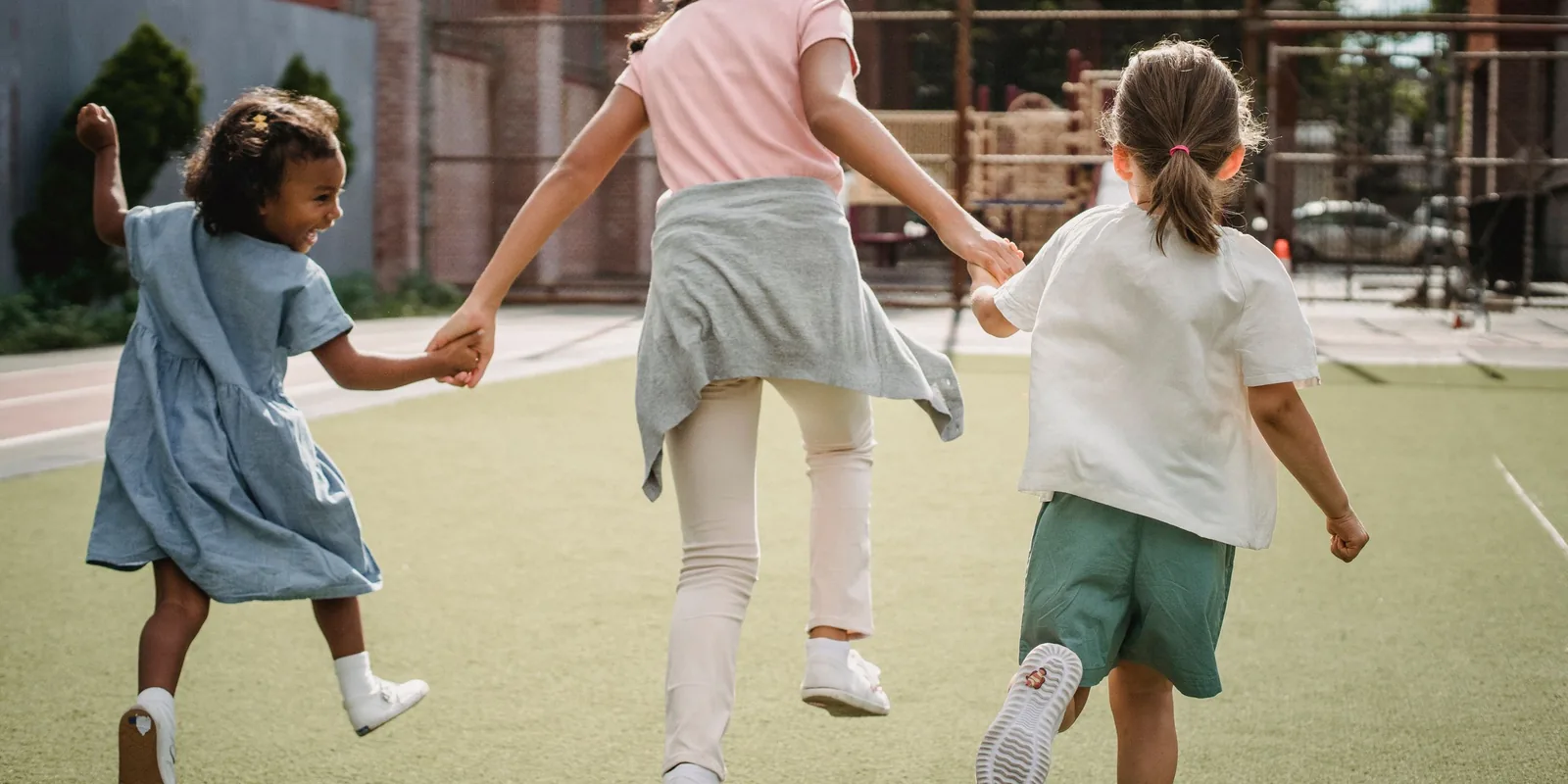 Three girls run hand in hand across a school playing field, one laughing over her shoulder