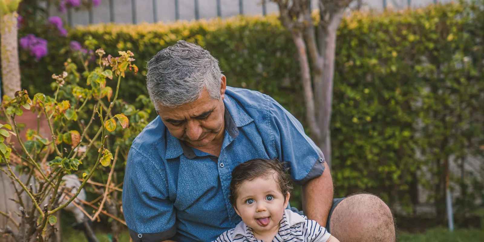 A grandfather crouches in a garden behind his baby grandson, who is sticking his tongue out