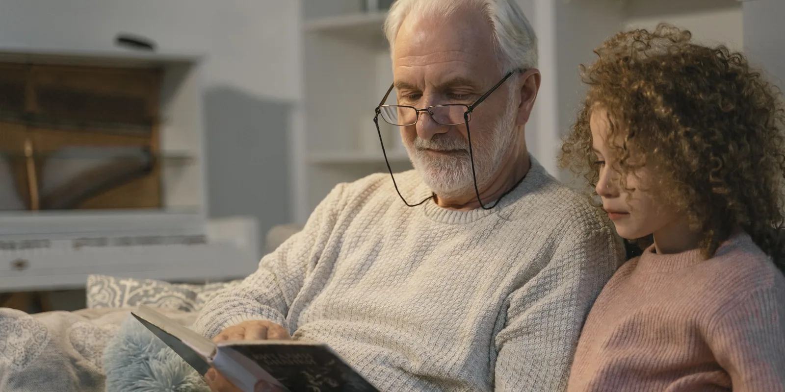 A white-haired man in reading glasses shares a book with a curly-haired girl on the sofa