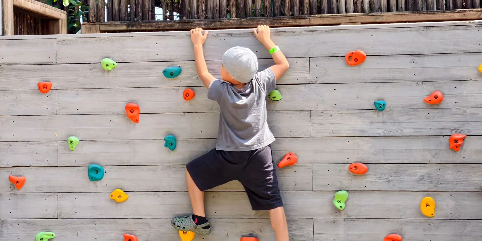 A boy in a cap reaches for the top of an outdoor climbing wall dotted with coloured holds