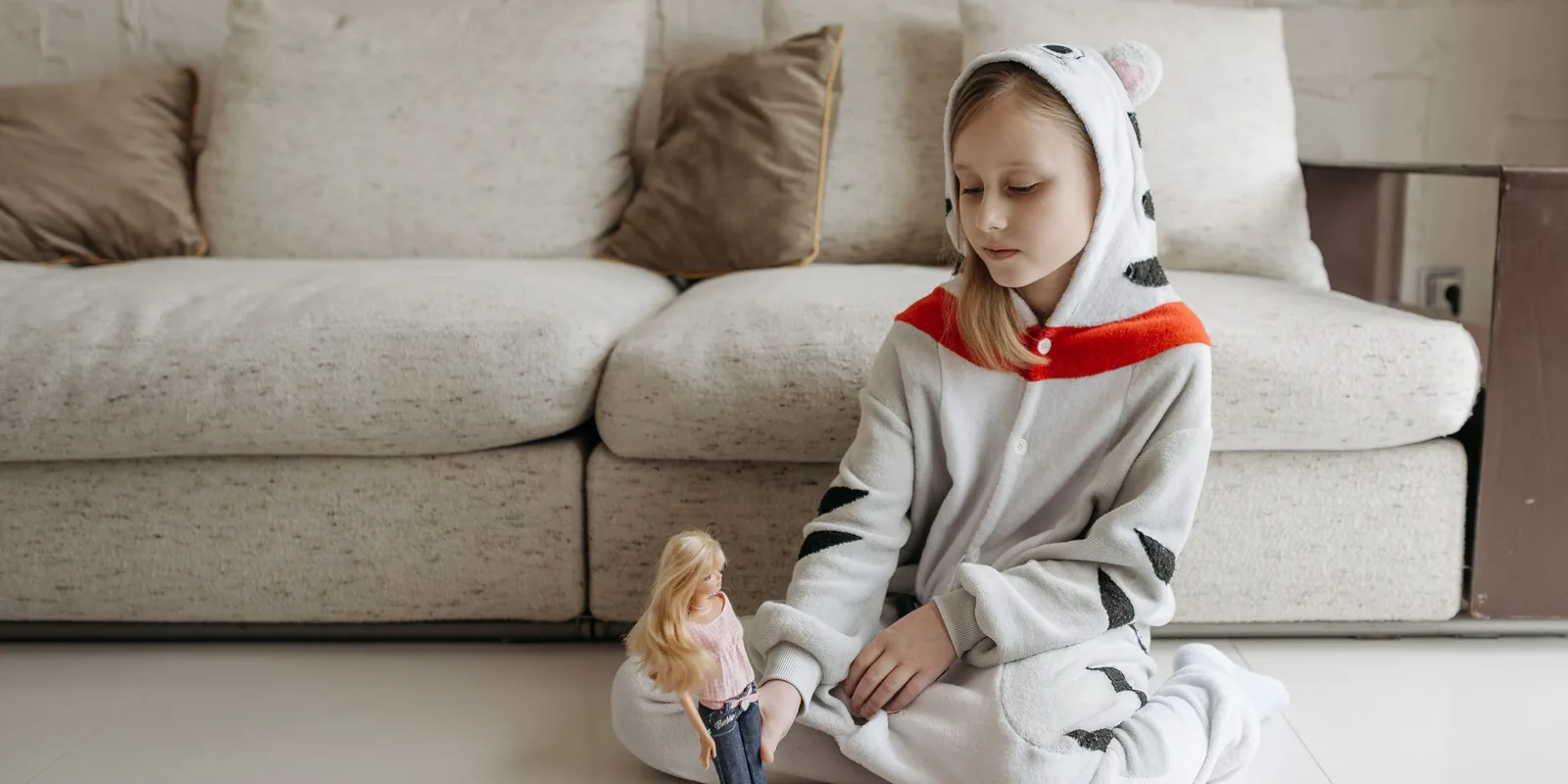 A girl in an animal onesie sits on the floor by a sofa playing with a doll on her own