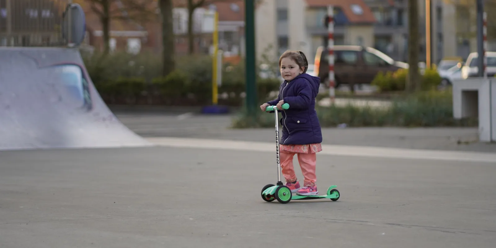 A small girl in a padded coat rides a green three-wheeled scooter across a skate park