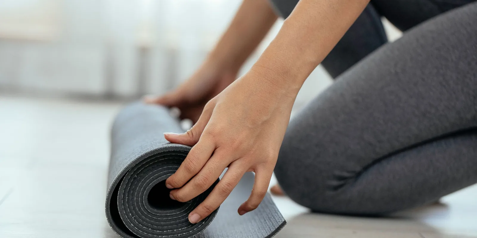 Close-up of hands rolling up a grey yoga mat on a pale wooden floor