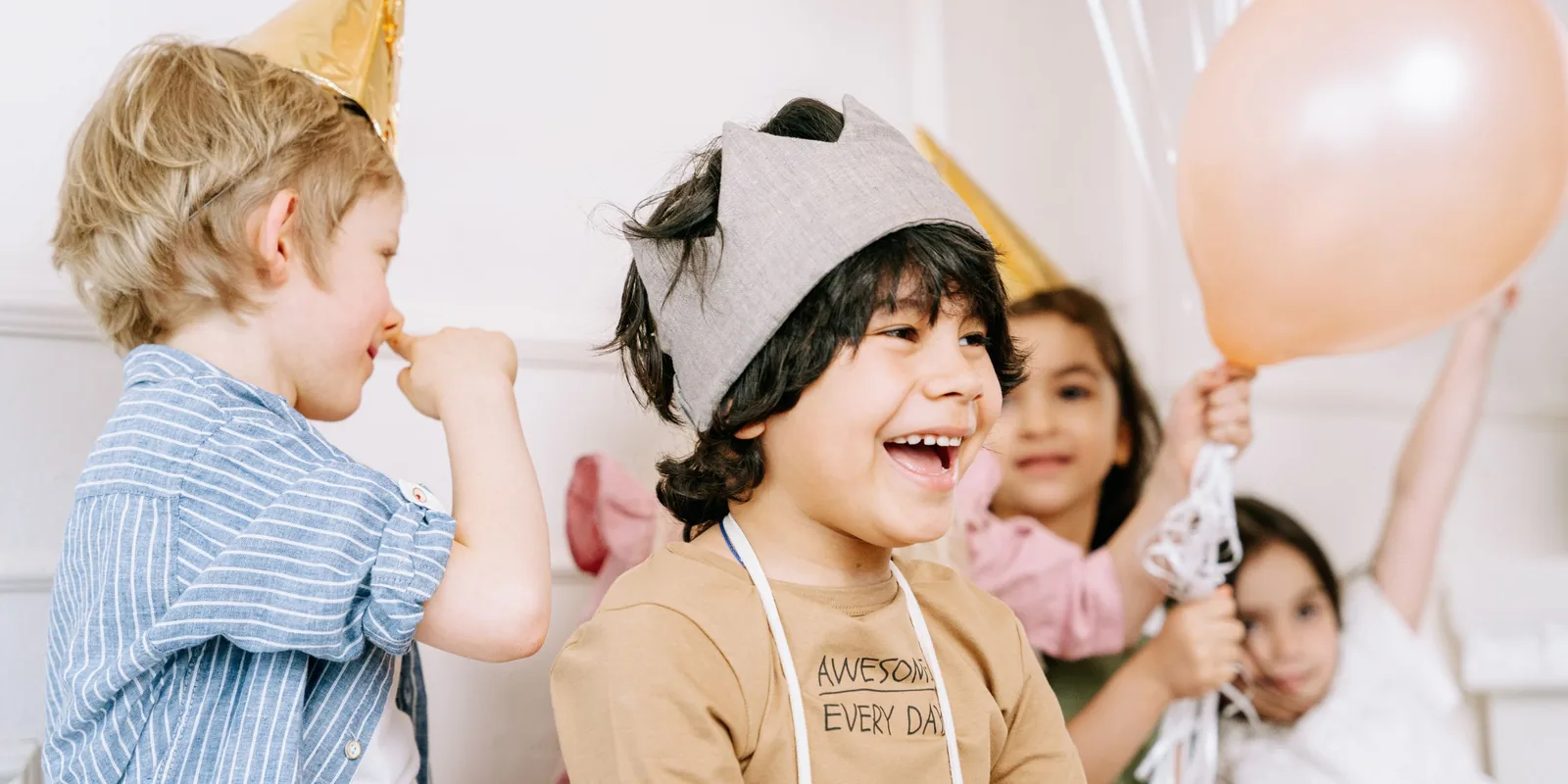 A laughing boy in a fabric crown stands among friends in party hats, one holding up a balloon