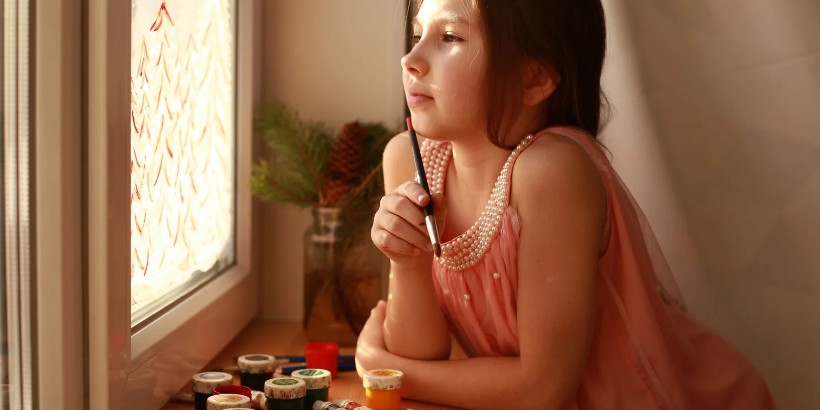 A girl gazes out of a sunny window, paintbrush resting on her chin, pots of paint on the sill