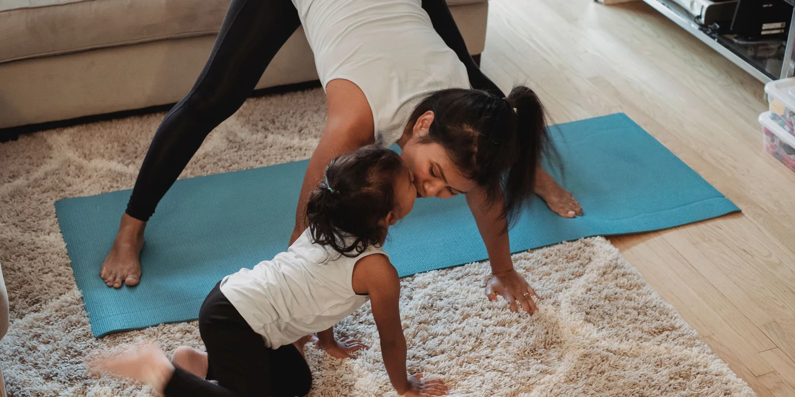 A woman in a yoga pose on a blue mat leans down to kiss a toddler crawling on the rug beside her