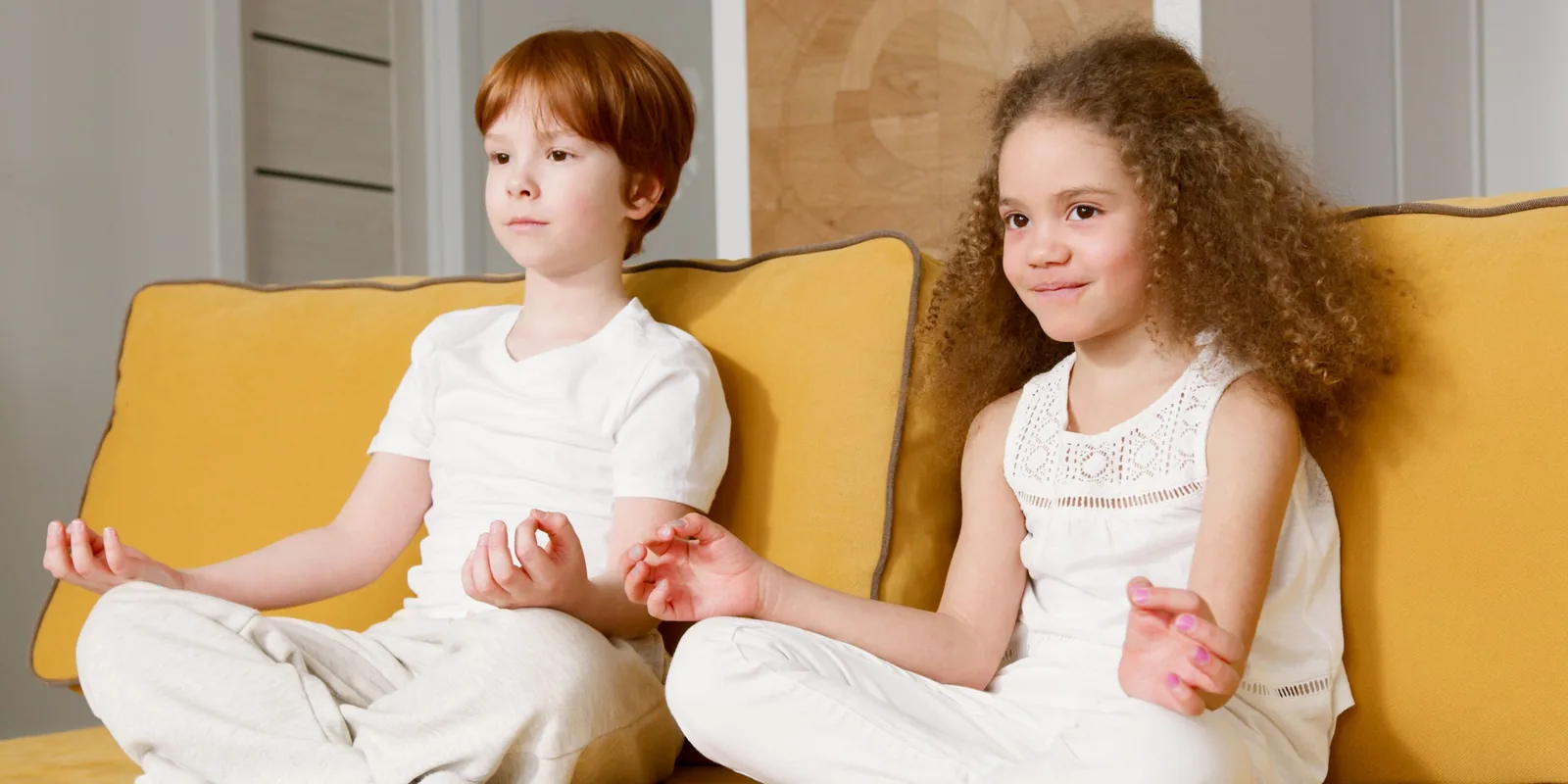 A boy and a girl dressed in white sit cross-legged on a yellow sofa, thumbs and fingers touching in a yoga meditation pose