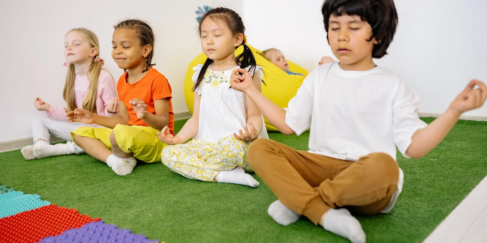 Four children sit cross-legged on a green floor with eyes closed and fingers in a meditation pose