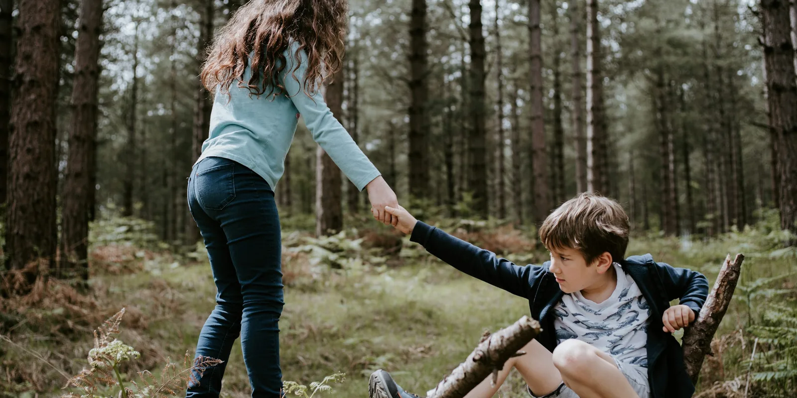In a pine wood, a girl reaches back to pull a boy up from where he has fallen among the branches