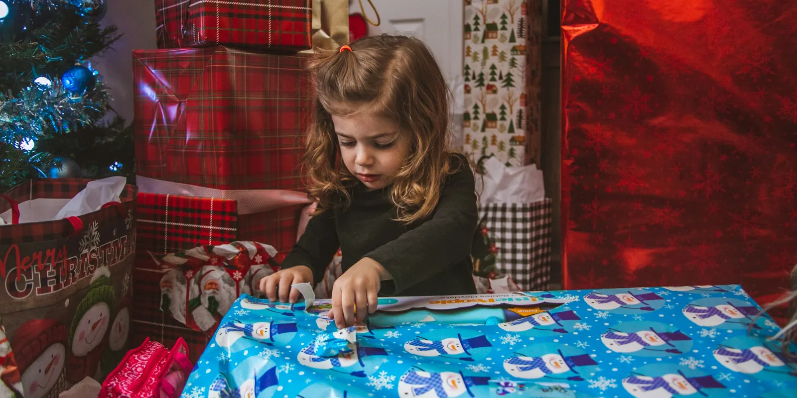 A young girl wraps a present in snowman paper beside a Christmas tree